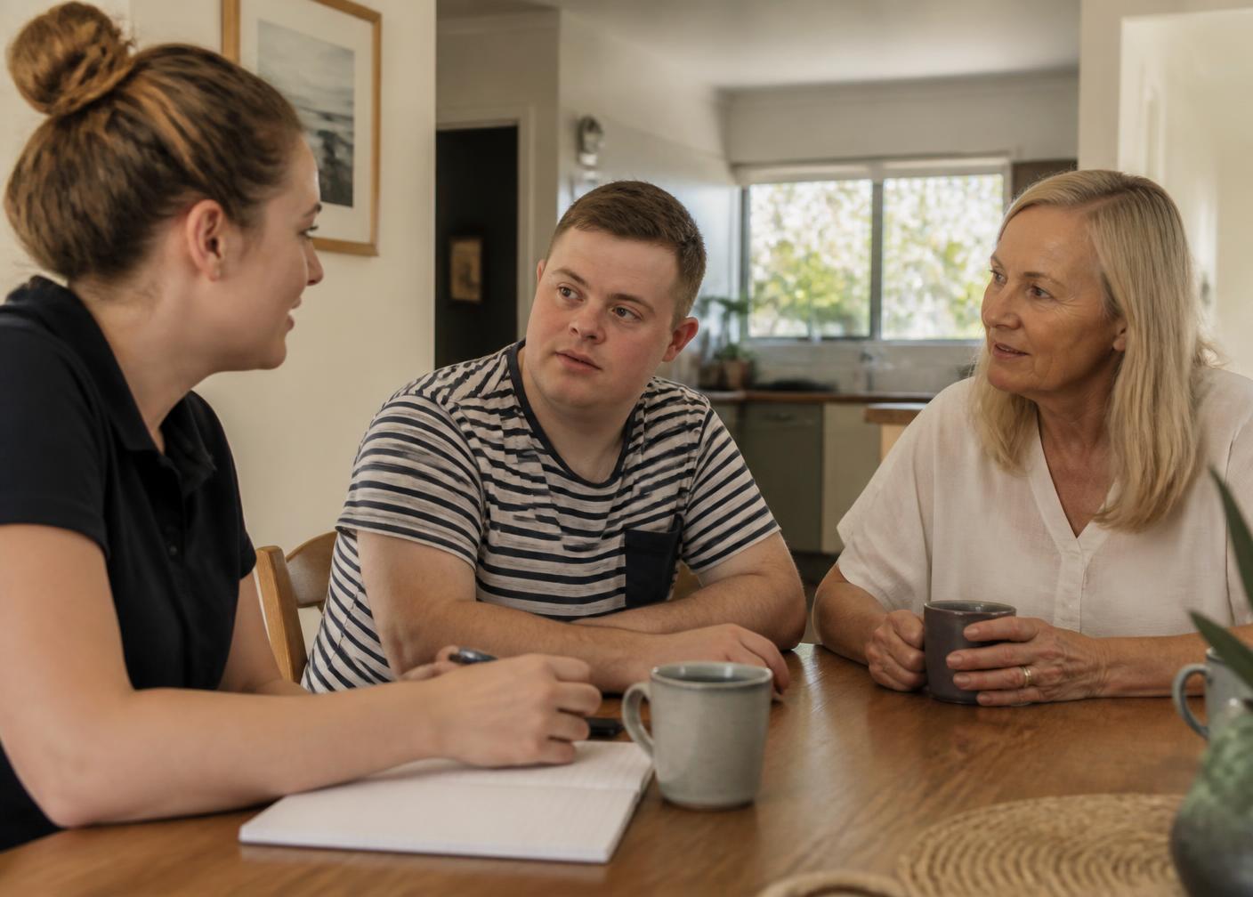 A support worker, a participant and a family member talking together around a kitchen table over cups of tea.