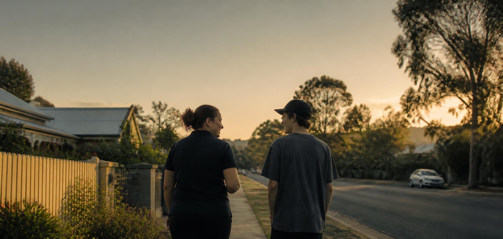 A support worker and a young man walking and talking along a leafy suburban street at sunset.